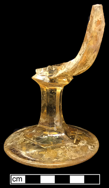 Colorless leaded stemmed glass.  Six-sided stem and panelled bowl.  This stemmed glass may have been a commercial container initially containing jelly and meant to be re-used as a goblet. Mold seams apparent on base (see detail photo). 3.25” base diameter. Yellowish color of vessel believed to be due to post-depositional causes.Appears similar to early 1870s examples from the King Company (Jones 2000:218).